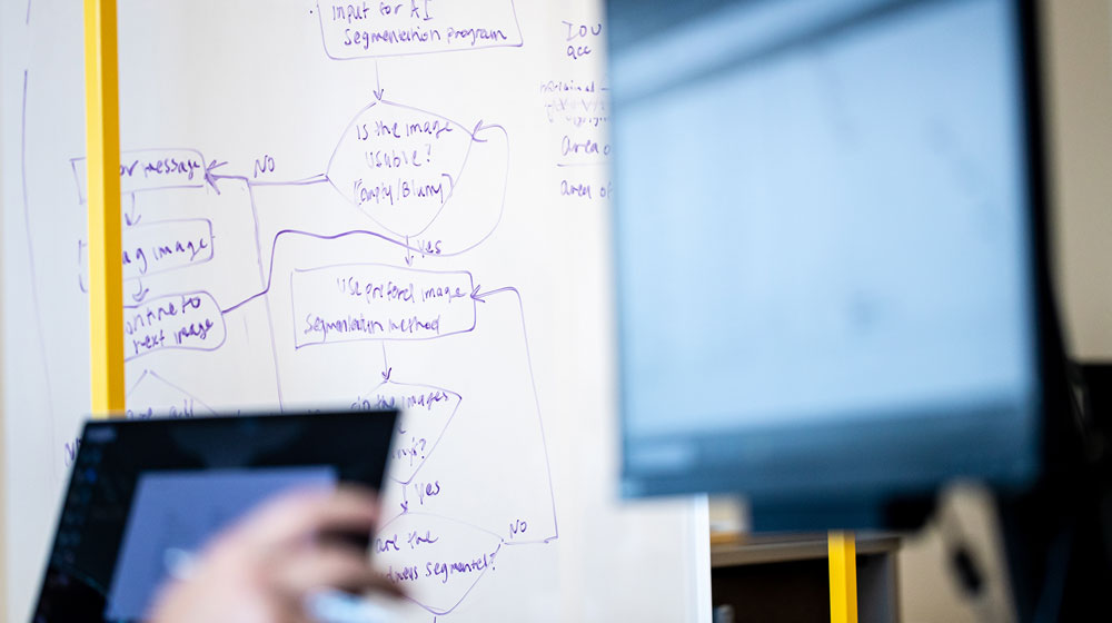 A diagram workflow is present on a whiteboard while an instructor teaches using classroom technology.