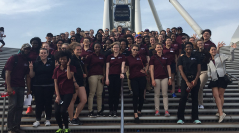 A large group of people in maroon shirts gather for a group photo on a large set a stairs outside