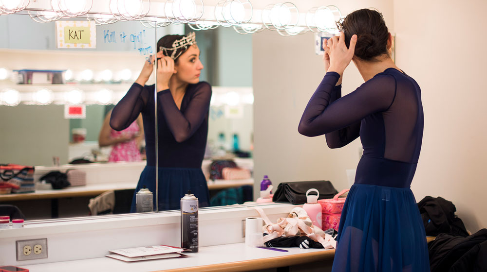 A dancer, in a black leotard, preparing for a performance, looks into a mirror in the dressing room as she sets her hair.