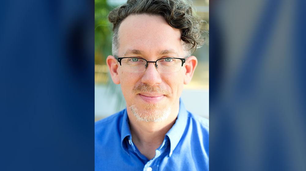 A portrait of faculty member Kevin McGroarty dressed in a blue button down shirt.He also wears wire-framed glasses.