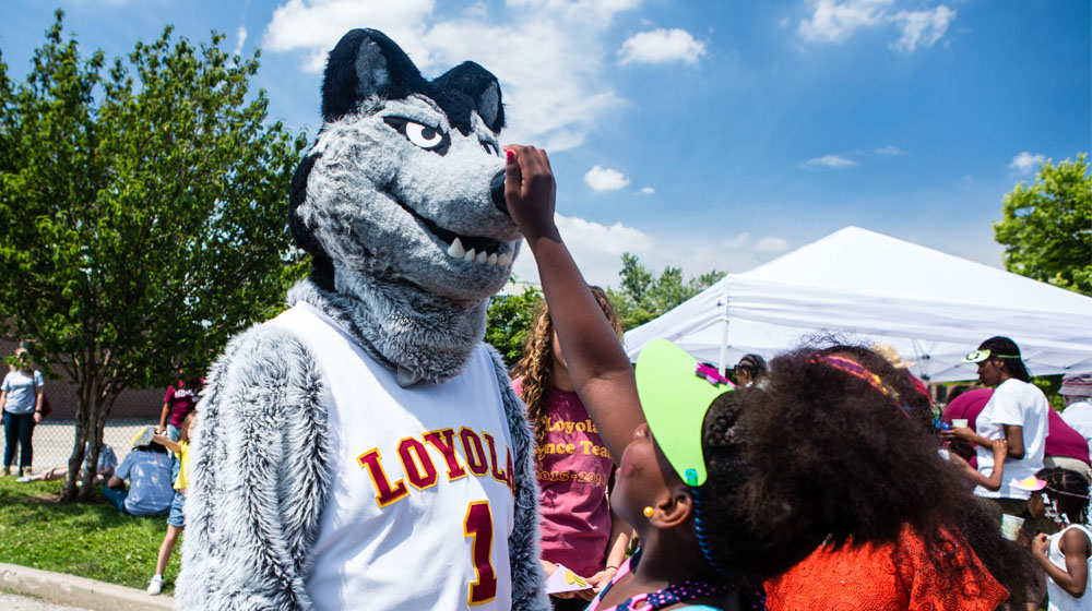 A young girl reaches up and touches the nose of the LU Wolf mascot costume outside with a blue sky and tress behind them.