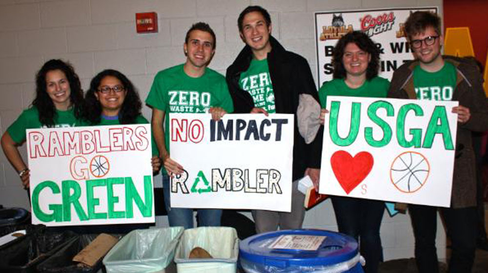 Six Loyola University Chicago students hold up hand made signs depicting their sustainability efforts for Zero Waste Game in front of recycling bins