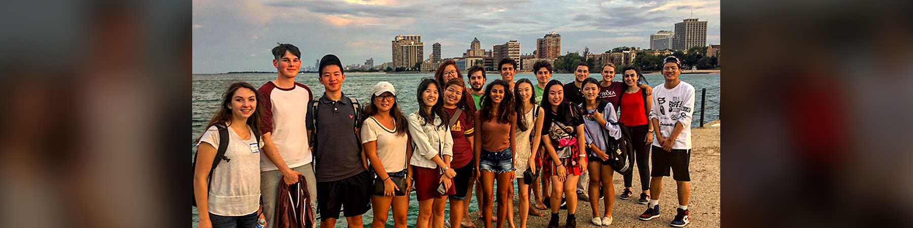 Diverse group of nearly twenty Loyola University Chicago international exchange students. Full body picture with everyone standing in a line on the walkway at the edge of Lake Michigan. The city of Chicago skyline is in the background on a partly cloudy day.