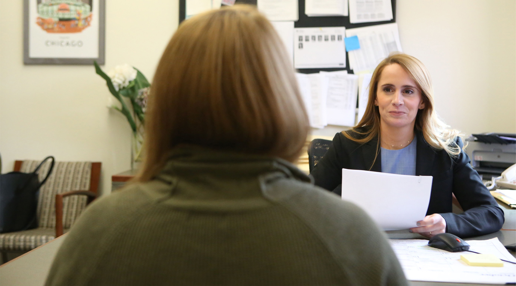 Emily Vaughn sits behind a desk, holding a paper. The back of a woman's head is visible in the foreground sitting on the other side of the desk.