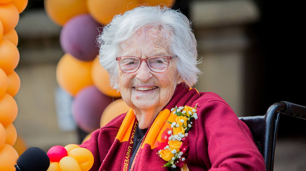 Sister Jean wears a maroon blazer with a five yellow rose corsage on the label and a striped maroon and gold scarf draped over the back of her neck.