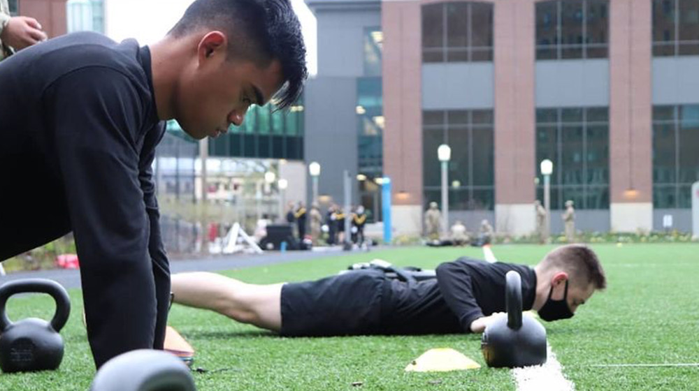 Two ROTC cadets in workout attire are doing pushups on a field. There are kettlebells near their hands.