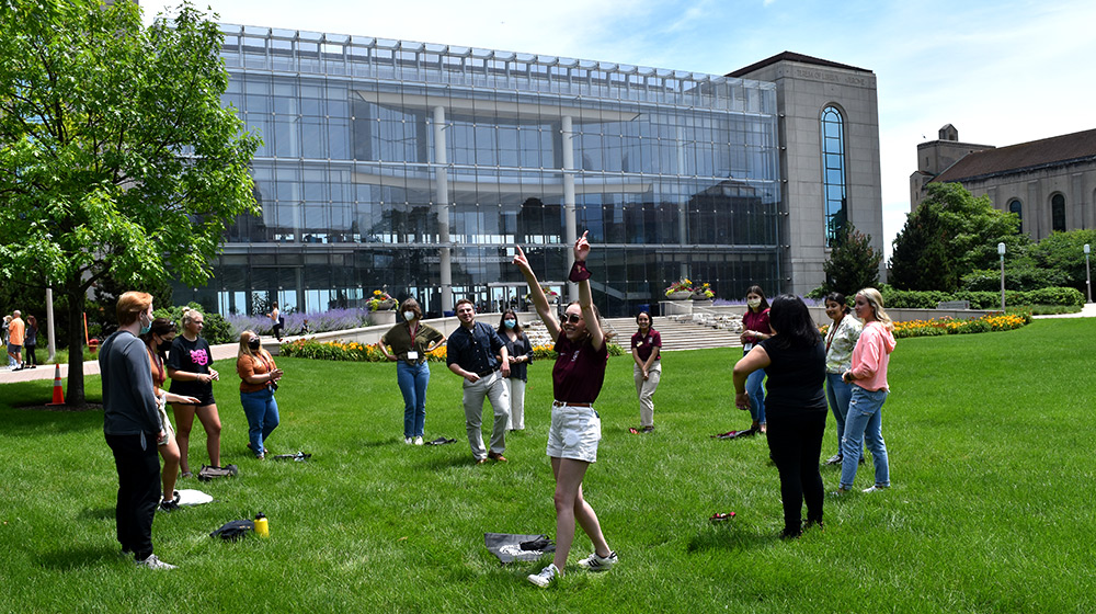 Multiple Loyola students gather in a circle outside on a grassy quad in front of a glass walled building.