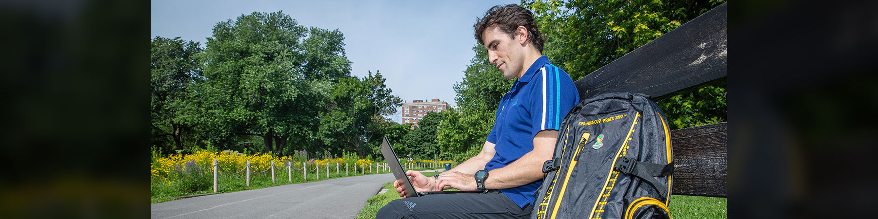 Loyola University Chicago white male student in a blue shirt, sitting on a bench typing on a laptop computer, accessing his Loyola courses online. His backpack is next to him on a sunny day with green trees and yellow flowers blooming along a bike path. There is a tall building in the distance. 