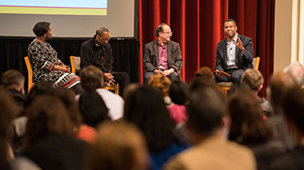 Four people sit on a stage discussing in front of an audience. The speaker on the right gestures while the others listen attentively. A large screen and red curtains are in the background. Audience members are blurred in the foreground.