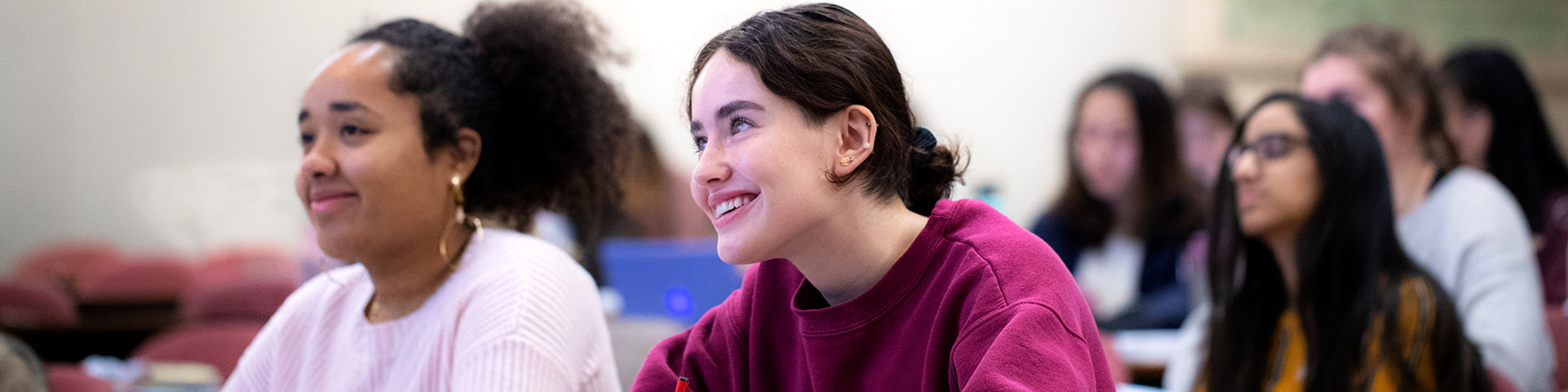 Multiple Loyola University Chicago students attend a sociology lecture. Two students in front are in focus while smiling and taking notes on the class while the rest are blurred in the background.