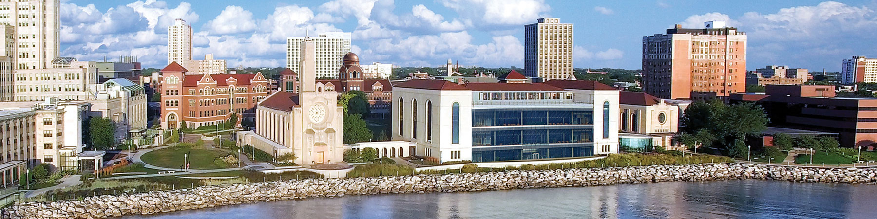 An aerial view of the Lake Shore Campus from lakeside with beautiful cumulus clouds in the sky.